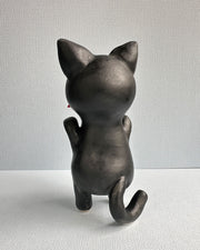 Ceramic sculpture of a black cat with large shiny eyes and pointed ears with pink interiors. It stands on its hind legs and has its paws up in the air and holds a red fish in its mouth.