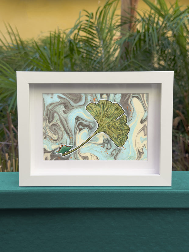 Illustration of a gingko leaf with a very small cartoon dinosaur at the bottom of it. A white outline lines the leaf and dinosaur, against a background of blue, gray and cream colored marbled paper. Piece is framed.