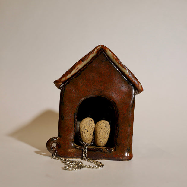 Ceramic sculpture of a brown dog house and a small character attached to it by a chain. The character is laying down within the house, only its feet are showing.