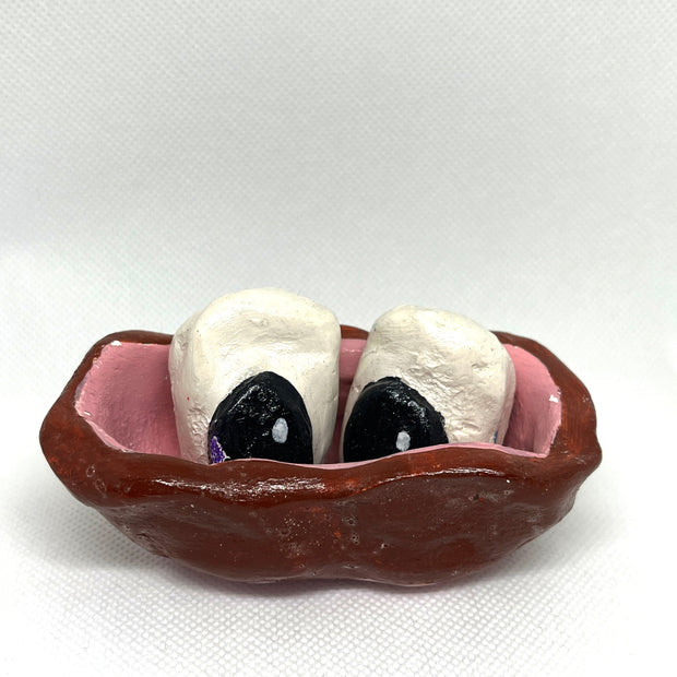 Clay sculpture of a stylistically messy brown bowl with 2 white ovals inside that are painted with black on them to look like eyes.