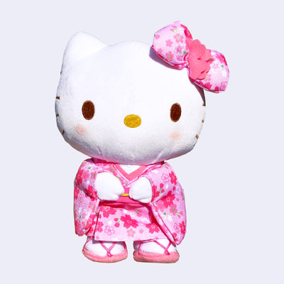 Plush doll of Hello Kitty, dressed in a pink kimono with cherry blossom pattern. She has a matching hair bow and sandals.