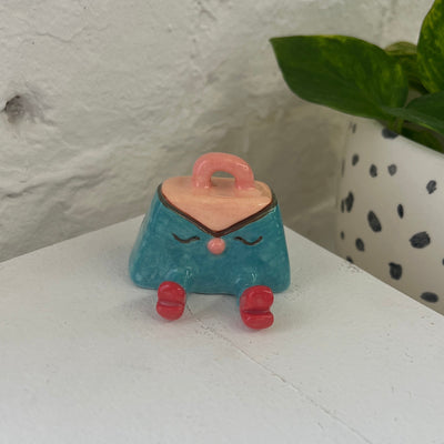 Small, shiny ceramic sculpture of a teal handbag with a pink handle. It has a simple closed eye expression with a small nose and a pair of legs. It sits and wears red shoes.