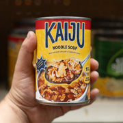 A stylized soup can being held in someone's hand. The can reads "Kaiju Noodle Soup" and has a photograph of a spoonful of brown soup with kaiju shaped noodles, over its bowl.