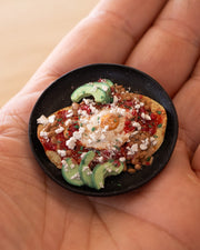 Small assembled clay sculpture of a model breakfast on a plate of huevos rancheros: eggs atop of tortillas with avocado, pico de gallo, pinto beans and cotija cheese. Piece held in the hand of the artist.