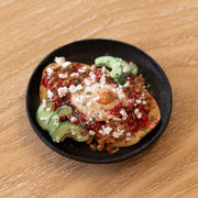 Small assembled clay sculpture of a model breakfast on a plate of huevos rancheros: eggs atop of tortillas with avocado, pico de gallo, pinto beans and cotija cheese.