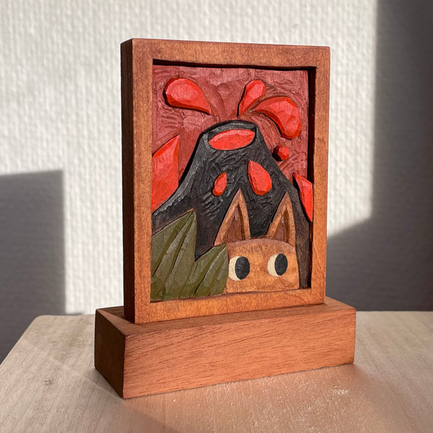 Painted relief carving into a rectangle of wood, depicting a semi 3D scene of an erupting volcano and a small creature viewing in the foreground, looking off to the side behind some leaves.