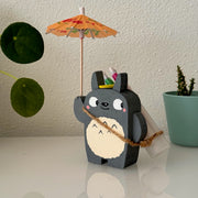  Small wooden sculpture of a smiling Totoro, looking off to the side. He holds up a small cocktail umbrella, with an over the shoulder bag holding 4 more umbrellas.