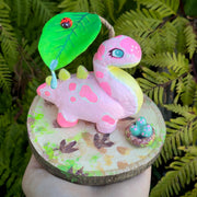 Sculpture on a circular wooden disk of a pink dinosaur with a long neck and spikes on its back. It has a cute face and a leaf hangs over its head with a water drip and a ladybug sitting on it. A small basket of eggs is nearby.