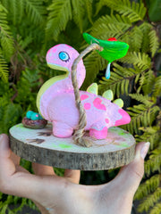 Sculpture on a circular wooden disk of a pink dinosaur with a long neck and spikes on its back. It has a cute face and a leaf hangs over its head with a water drip and a ladybug sitting on it. A small basket of eggs is nearby.