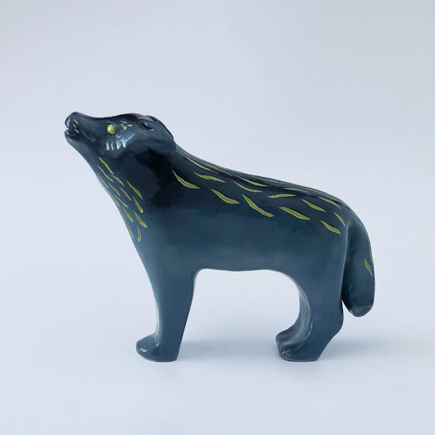 Ceramic sculpture of a bluish black wolf, standing and howling at the moon. Its two pairs of legs (front and back respectively) are paired together and it has gold eyes and fur color accents.