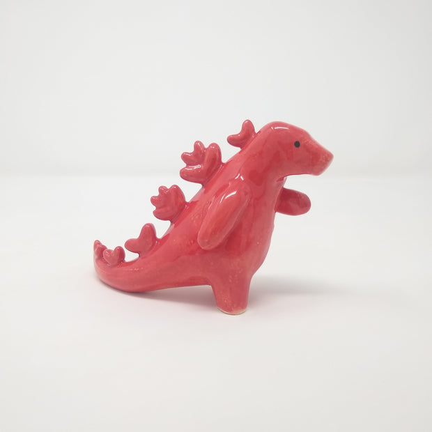 Glazed ceramic sculpture of a red Godzilla like moster with rounded spikes on its back and one hand extended out. It has simple drawn on eyes and no other facial features.