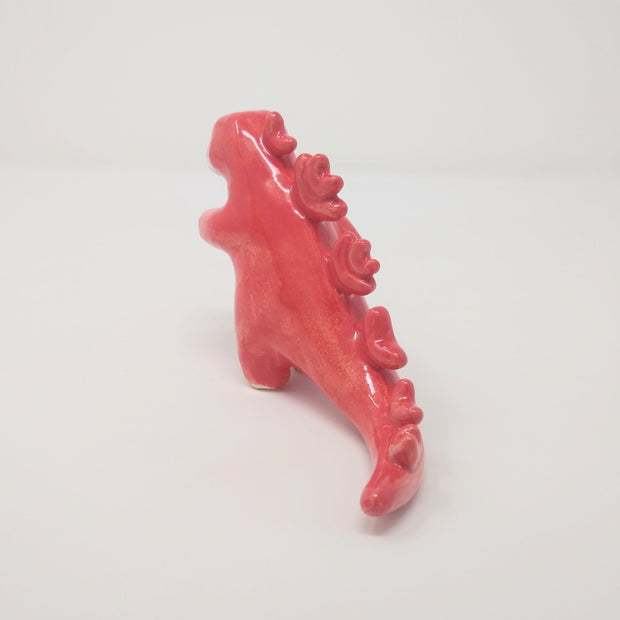 Glazed ceramic sculpture of a red Godzilla like moster with rounded spikes on its back and one hand extended out. It has simple drawn on eyes and no other facial features.