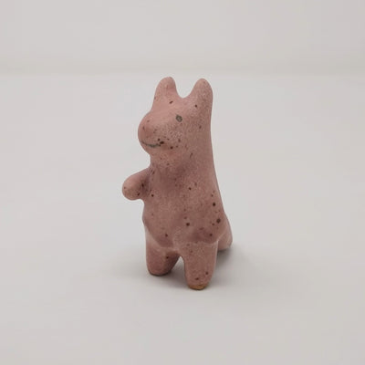 Ceramic sculpture of a brown devil like character with rounded horns and simple rounded limbs. It stands with one hand extended and a simple drawn on smiling face.