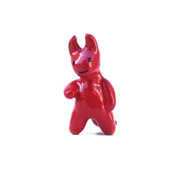 Glazed ceramic sculpture of a red devil with soft edged horns and a simple drawn on smile. It has rounded limbs and extends one arm out.