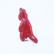 Glazed ceramic sculpture of a red devil with soft edged horns and a simple drawn on smile. It has rounded limbs and extends one arm out.