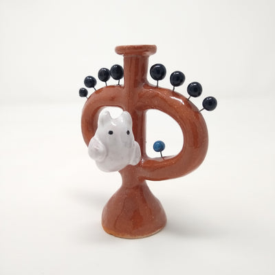  Ceramic sculpture of a brown vessel with handles and a small flat area up top. A white ghost with small devil horns is on the front of the sculpture. Small black balls come out of the vessel via wire.