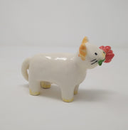 Small ceramic sculpture of a white cat with yellow ears and paws and 2 tails. It holds a red flower in its mouth.