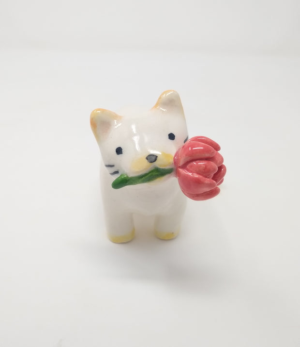 Small ceramic sculpture of a white cat with yellow ears and paws and 2 tails. It holds a red flower in its mouth.