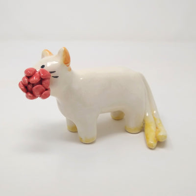 Small ceramic sculpture of a white cat with yellow ears and paws and 2 tails. It holds a red flower in its mouth.