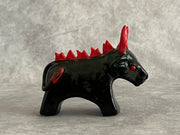 Small black ceramic sculpture of a bull with an angry expression, red eyes, red horns and a red mane on its back. It stands with its front legs fused together into one unit, and its back legs fused into their own unit.