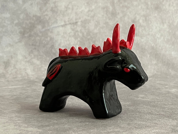 Small black ceramic sculpture of a bull with an angry expression, red eyes, red horns and a red mane on its back. It stands with its front legs fused together into one unit, and its back legs fused into their own unit.