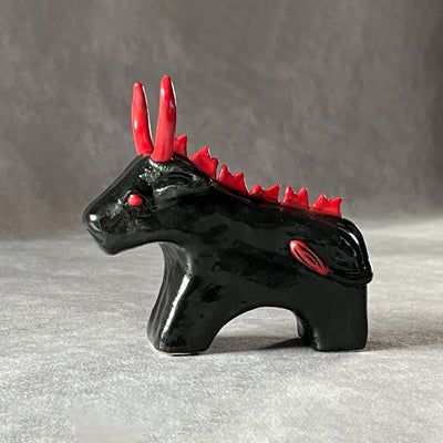 Small black ceramic sculpture of a bull with an angry expression, red eyes, red horns and a red mane on its back. It stands with its front legs fused together into one unit, and its back legs fused into their own unit.