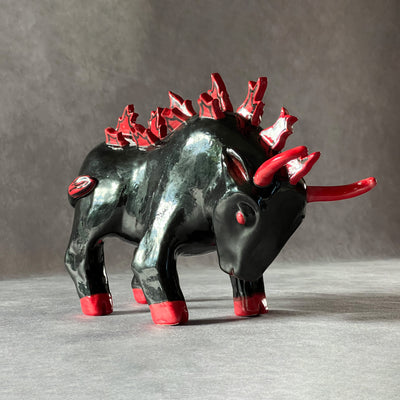 Black ceramic sculpture of a bull, faced down with an angry expression and red color accents for its eyes, horns, hooves and mane. Atop its back are red flames in place of a mane.