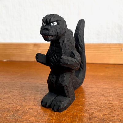 Whittled wooden sculpture of a black Godzilla character, with white eyes and a stern expression. It stands with its arms slightly raised at its sides and its large tail behind it.