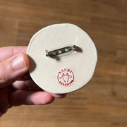 Back side of ceramic circular brooch with a metal brooch backing attached and the artist's stamped signature.