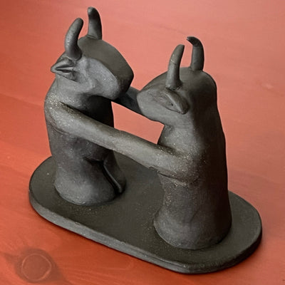 Black matte ceramic sculpture of 2 figures with bull heads, standing and facing one another. Their arms are extended out and connect with each other seamlessly, creating one long line from one figure to the other.