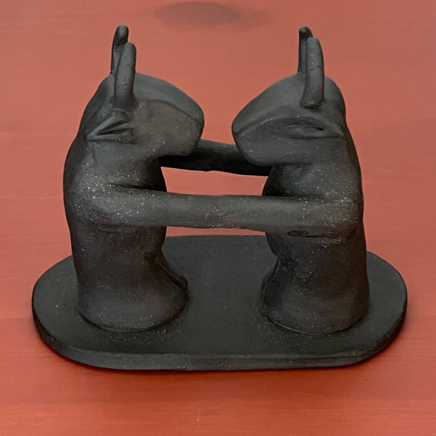 Black matte ceramic sculpture of 2 figures with bull heads, standing and facing one another. Their arms are extended out and connect with each other seamlessly, creating one long line from one figure to the other.