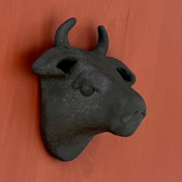 Black matte clay sculpture of a bull's head mounted onto the wall and flat on the back.