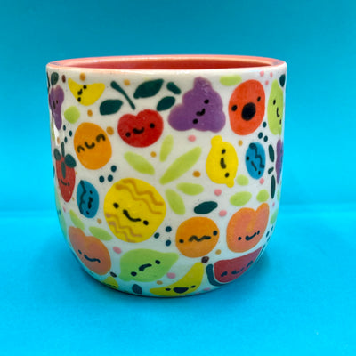 Ceramic cup covered with cute, cartoon illustrations of very colorful fruit with simple cartoon faces. Fruits include: grapes, cherries, citrus, pineapple, watermelon, etc.