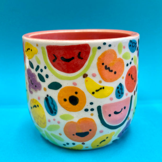 Ceramic cup covered with cute, cartoon illustrations of very colorful fruit with simple cartoon faces. Fruits include: grapes, cherries, citrus, pineapple, watermelon, etc.
