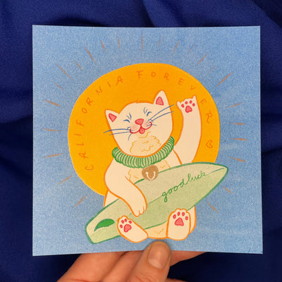 Risograph print of a white cat, styled like a Japanese lucky cat with a green collar and a bell on its neck. It holds a small green surfboard that reads "good luck".