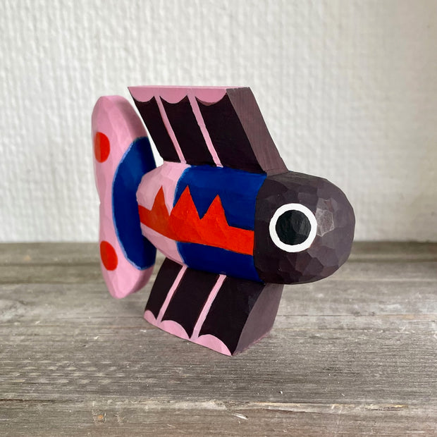 Brightly painted whittled wooden sculpture of a fish colored black with blue and pink body and tail and red accent coloring. It has a set of eyes, one on each side of its head.