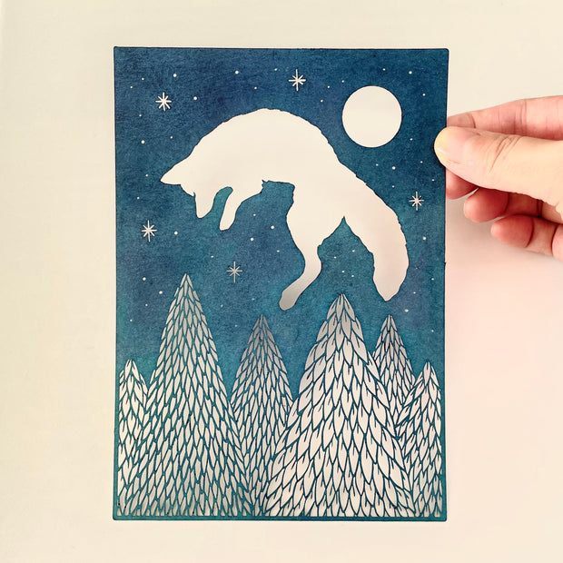 Blue paper with cut out imagery of a jumping fox, which leaps over a row of cut out pine trees. In the top right corner is a full moon and smaller stars.