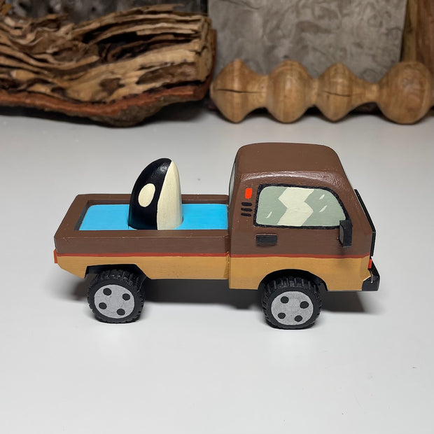 Painted small whittled wooden sculpture of a brown striped pick up truck, with the pick up filled with water. The head of a killer whale peeks out from the water.