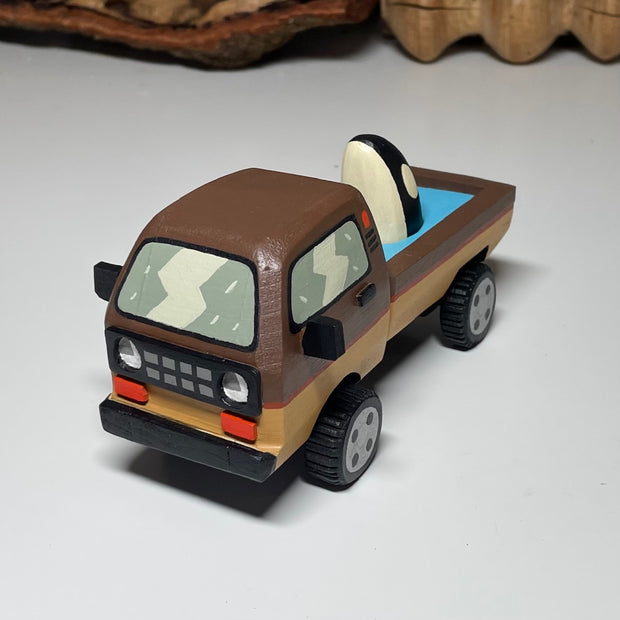 Painted small whittled wooden sculpture of a brown striped pick up truck, with the pick up filled with water. The head of a killer whale peeks out from the water.