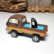 Painted small whittled wooden sculpture of a brown striped pick up truck, with the pick up filled with water. The head of a killer whale peeks out from the water.