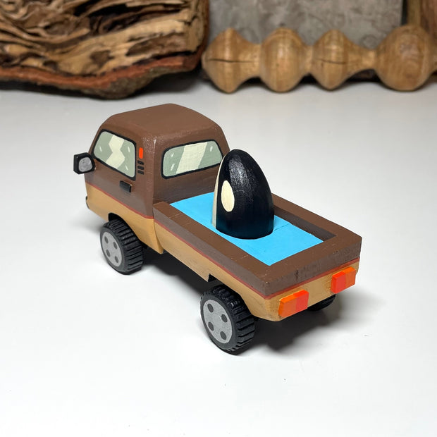 Painted small whittled wooden sculpture of a brown striped pick up truck, with the pick up filled with water. The head of a killer whale peeks out from the water.