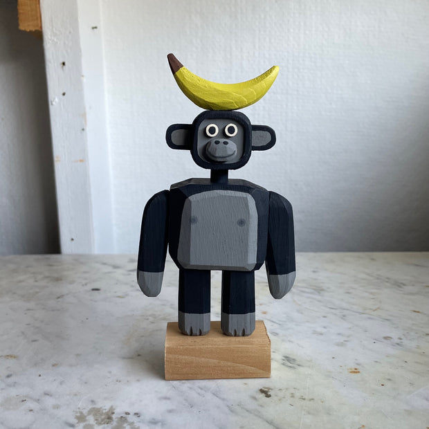 Assembled wooden sculpture of a smiling cartoon gorilla with a thin neck and thick arms and legs. Balancing atop its head is a banana.
