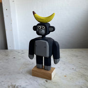 Assembled wooden sculpture of a smiling cartoon gorilla with a thin neck and thick arms and legs. Balancing atop its head is a banana.