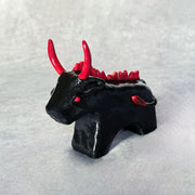 Small black ceramic sculpture of a bull with an angry expression, red eyes, red horns and a red mane on its back. It stands with its front legs fused together into one unit, and its back legs fused into their own unit.