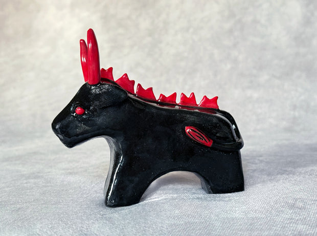 Small black ceramic sculpture of a bull with an angry expression, red eyes, red horns and a red mane on its back. It stands with its front legs fused together into one unit, and its back legs fused into their own unit.