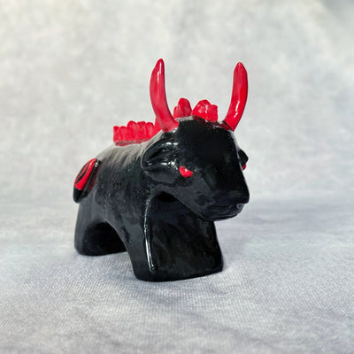 Small black ceramic sculpture of a bull with an angry expression, red eyes, red horns and a red mane on its back. It stands with its front legs fused together into one unit, and its back legs fused into their own unit.