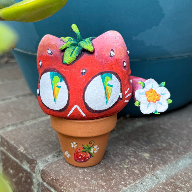 Sculpture in a small planter pot of a large cartoon cat head, made to look like a strawberry with large green and blue eyes. It has no limbs but a small white flower blooms from its tail.