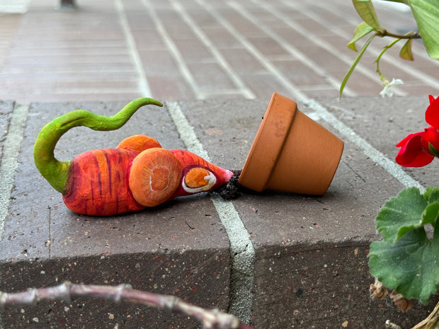 Small sculpture of a rat made to look like a carrot with its body and carrot slice ears. It has a green, vine like tail and sniffs through a spilled plant pot with a slice of cheese inside.