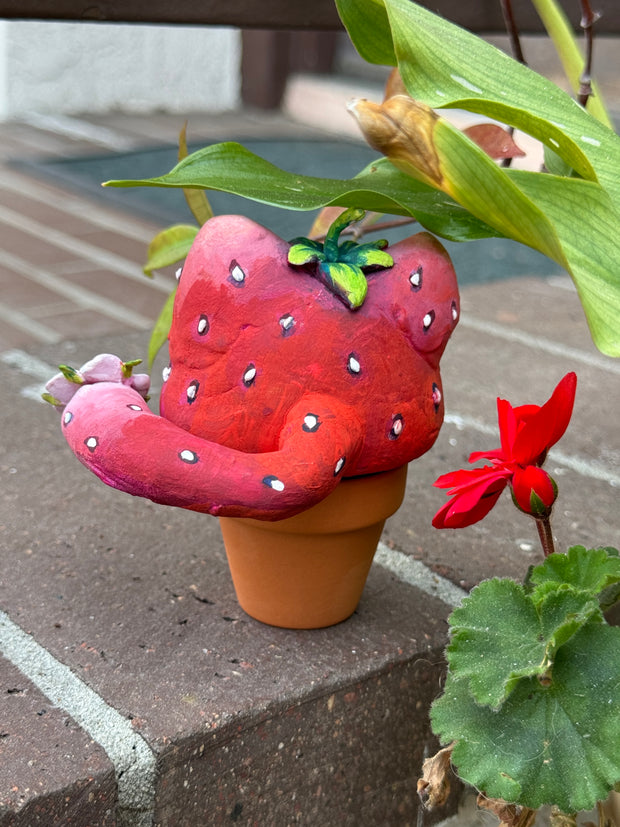 Sculpture in a small planter pot of a large cartoon cat head, made to look like a strawberry with large green and blue eyes. It has no limbs but a small white flower blooms from its tail. Backside.