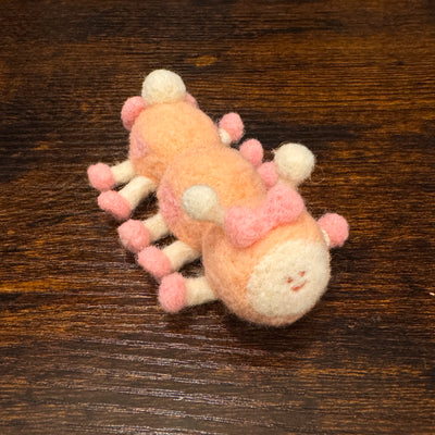 Felted sculpture of a pastel orange caterpillar with a small smiling face and 10 legs, all spread out flat so it is laying instead of standing. It wears pink shoes on all its feet and has a matching pink bow.